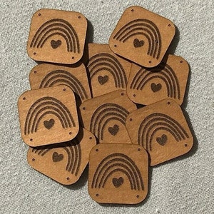 Könnte beinhalten: Eine Ansammlung kleiner, quadratischer Holzverzierungen. Jedes Stück hat ein lasergraviertes Design eines Regenbogens und eines Herzens. Das Holz hat eine natürliche, hellbraune Farbe und die Kanten sind leicht abgerundet. Kleine Löcher sind in jeder Ecke gebohrt.