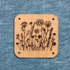 May include: A brown wooden tag with a laser-etched design of a field of wildflowers. The tag has rounded corners and a slightly rough texture.