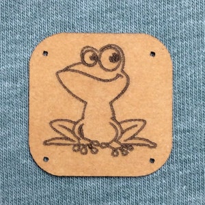 Può includere: Una toppa quadrata, color cuoio, con un'illustrazione di un ranocchio dei cartoni animati, cucita su un tessuto verde acqua. La rana è disegnata in uno stile semplice, con grandi occhi e un'espressione sorridente. La toppa ha quattro piccoli fori.