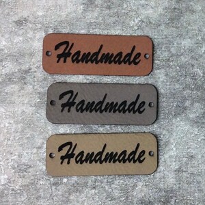 Può includere: Tre etichette rettangolari in pelle con la scritta "Handmade" in nero su ogni etichetta. Le etichette sono marrone, grigio e beige.
