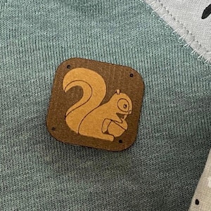Puede incluir: Un parche de tela marrón con un contorno marrón claro, que presenta una ardilla de dibujos animados sosteniendo una nuez. El parche está cosido sobre un fondo de tela verde.