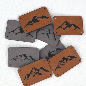 以下が含まれることがあります： 長方形の革製パッチのコレクション。グレーと茶色があります。各パッチには黒い山脈のデザインが施されています。パッチは角が丸く、縫い付けたり取り付けたりするための小さな穴があります。