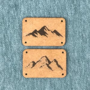 Könnte beinhalten: Zwei braune Holzetiketten mit einem schwarzen Bergsilhouetten-Design. Die Etiketten haben abgerundete Ecken und vier kleine Löcher.