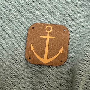 Puede incluir: Primer plano de una tela verde claro con un parche cuadrado marrón con un diseño de ancla beige. El parche está sujeto a la tela con pequeños remaches. Una cremallera blanca es visible en el lado derecho de la tela.