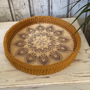 May include: A round wooden tray with a crocheted mustard yellow border. The tray features a laser-etched mandala design with a flower of life symbol in the center.