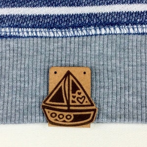 May include: Close-up of a blue and white striped sweatshirt with a gray collar and hem. A small, rectangular wooden tag with a sailboat design is attached to the hem. The sailboat has a heart and decorative details.