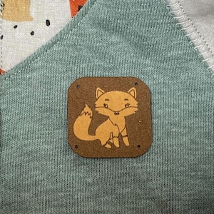 Puede incluir: Primer plano de una prenda de bebé con un panel verde y una cremallera. La prenda presenta un patrón repetido de animales del bosque sobre un fondo claro. Un pequeño parche cuadrado marrón con una ilustración de zorro está cosido en el panel verde.