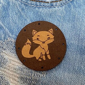 May include: A brown leather patch with a fox design sewn onto a light blue denim fabric. The fox is brown with a white chest and a white muzzle.