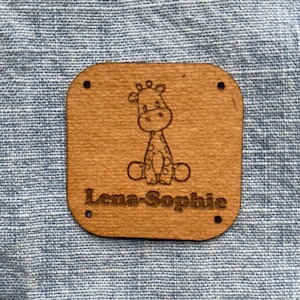 Puede incluir: Una etiqueta de cuero marrón con una ilustración de jirafa y el nombre "Lena-Sophie" grabado en ella.
