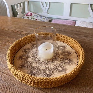 May include: A round wooden tray with a crocheted mustard yellow border. The tray has a mandala design burned into the wood. A glass candle holder with a white candle sits in the center of the tray.