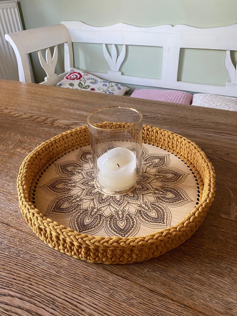 May include: A round wooden tray with a crocheted mustard yellow border. The tray has a mandala design burned into the wood. A glass candle holder with a white candle sits in the center of the tray.