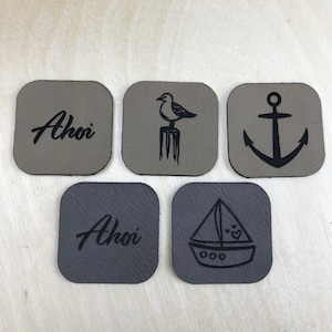 Könnte beinhalten: Fünf quadratische Leder-Patches mit verschiedenen Designs. Zwei Patches haben das Wort "Ahoy" in Kursivschrift. Ein Patch hat eine Möwe, die auf einem Pfosten steht. Ein Patch hat einen Anker. Ein Patch hat ein Segelboot mit Herzen.