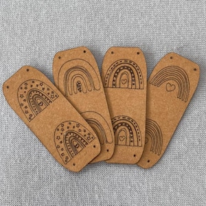 Puede incluir: Cinco recortes de arcoíris de madera sin terminar con diferentes diseños. Cada recorte tiene un corazón en el centro y dos agujeros en la parte superior.