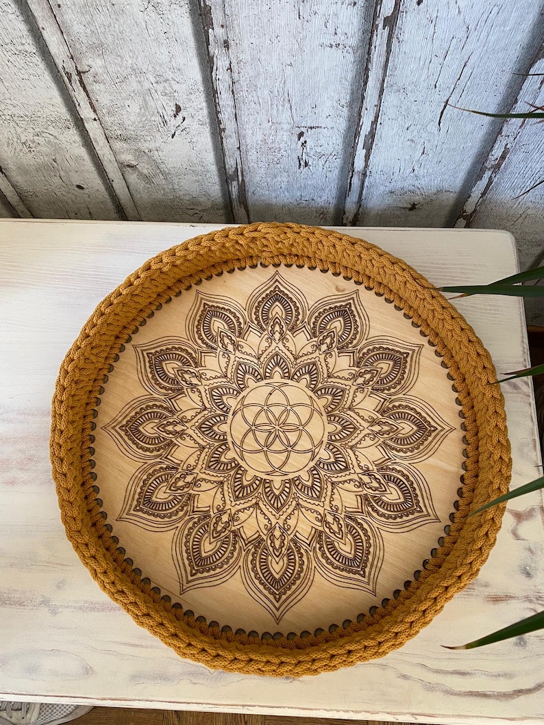 May include: A round wooden tray with a brown mandala design and a mustard yellow crocheted border.