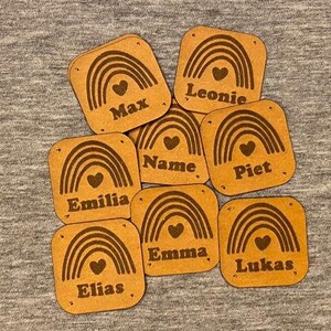 Puede incluir: Ocho parches de cuero marrón con diseños de arcoíris y nombres bordados. Los nombres son Max, Leonie, Name, Piet, Emilia, Emma, Elias y Lukas.