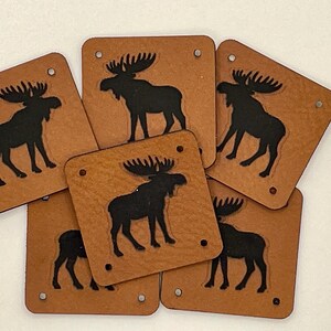 May include: Six square, brown leather coasters with a black moose silhouette. Each coaster has rounded corners and three small holes. The moose design is centered on each coaster.
