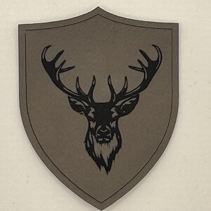 Pode incluir: Um patch cinza com um desenho de cabeça de veado preto. A cabeça do veado está centrada em um patch em forma de escudo. O patch tem uma borda cinza escuro.
