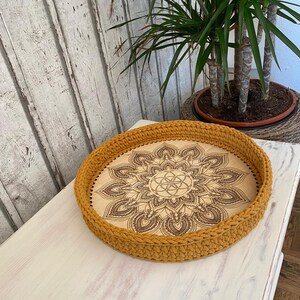 May include: A round wooden tray with a crocheted mustard yellow border. The tray features a laser-etched mandala design with a geometric pattern in the center.