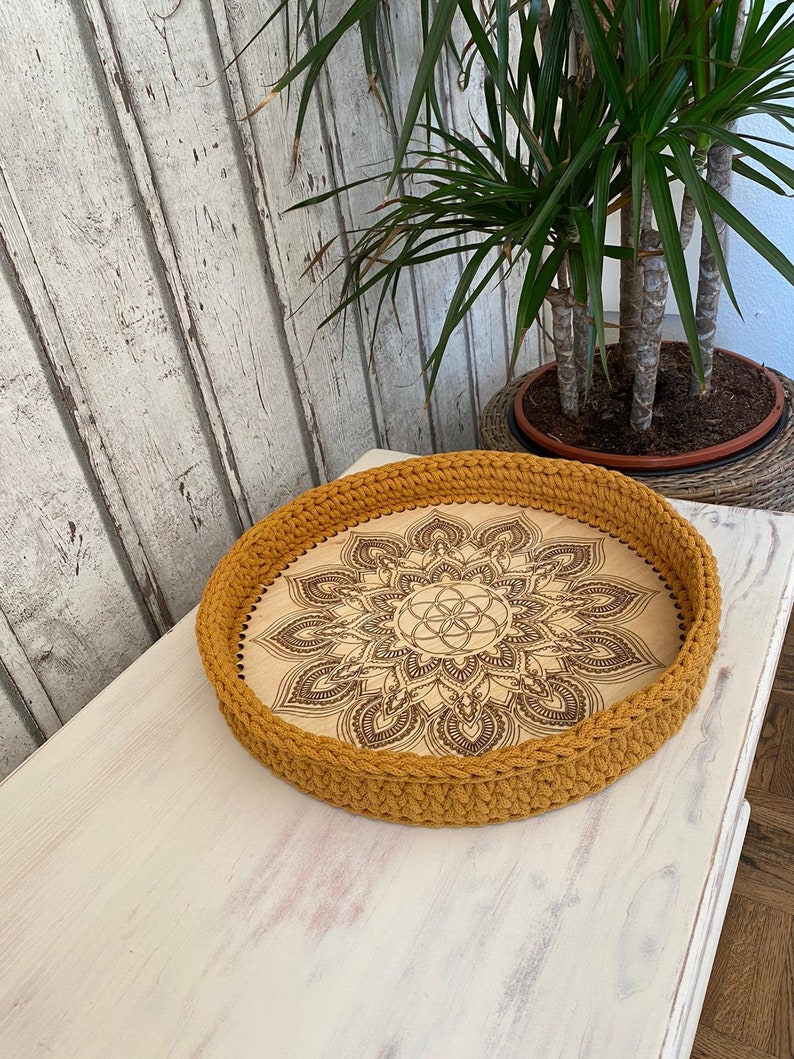 May include: A round wooden tray with a crocheted mustard yellow border. The tray features a laser-etched mandala design with a geometric pattern in the center.