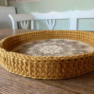 May include: A round, mustard yellow crocheted tray with a wooden base featuring a mandala design.