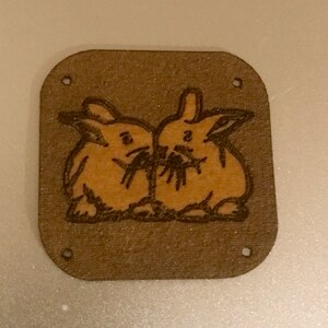 May include: A brown wooden square with two brown rabbits drawn on it. The rabbits are facing each other and appear to be eating something.