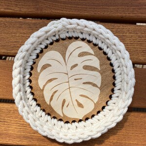 Könnte beinhalten: Ein weißer gehäkelter Untersetzer mit einem runden Holzeinsatz, der ein lasergeschnittenes weißes Monstera-Blattdesign zeigt.