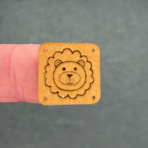 Puede incluir: Pequeño adorno cuadrado de madera con la cara de un león y una melena ondulada. La cara del león es un simple dibujo lineal con una expresión sonriente. El adorno tiene pequeños agujeros en cada esquina.