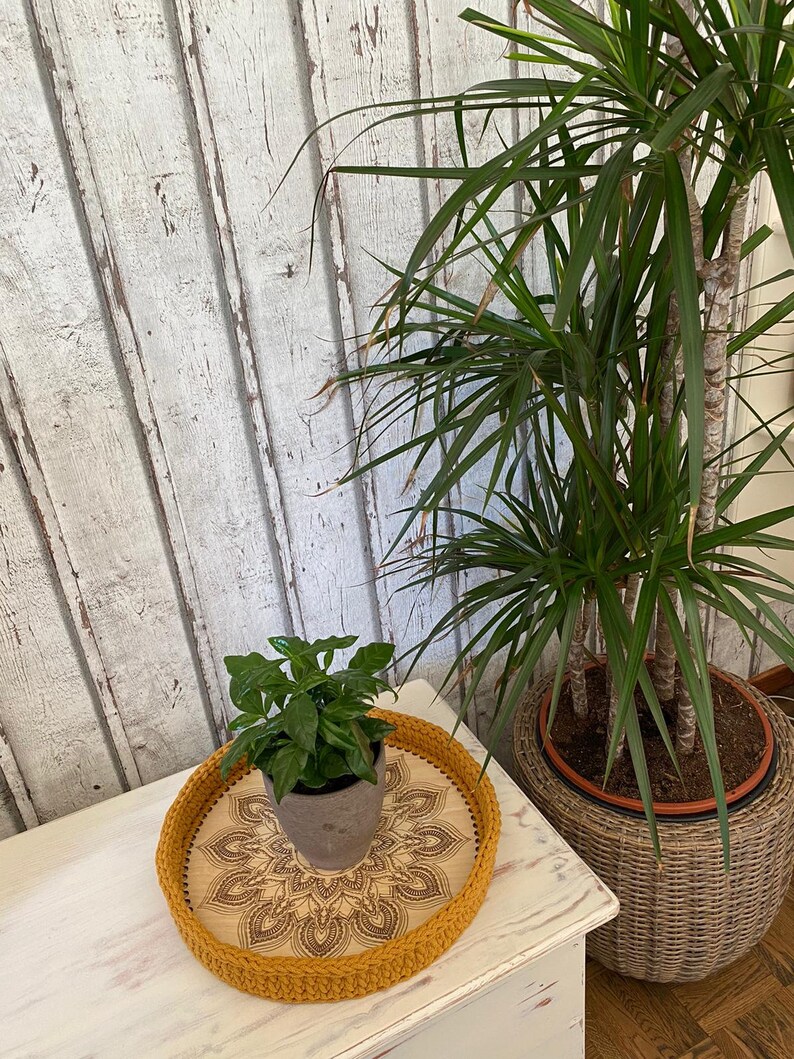 May include: A yellow crocheted tray with a mandala design on the bottom. The tray is on a white wooden surface and has a small potted plant inside.