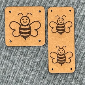 May include: Two brown rectangular tags with a cute bee illustration. The bee is smiling with striped body and wings. The tags have small holes in each corner. The tags are on a gray fabric background.