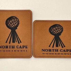 Puede incluir: Dos parches de cuero marrón con esquinas redondeadas. Cada parche presenta un gráfico negro de un globo terráqueo sobre una base de trípode, con el texto "NORTH CAPE" y coordenadas debajo. Los parches son de diferentes tamaños.
