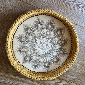 May include: A round wooden tray with a crocheted mustard yellow border. The tray features a laser-etched mandala design with a flower of life in the center.