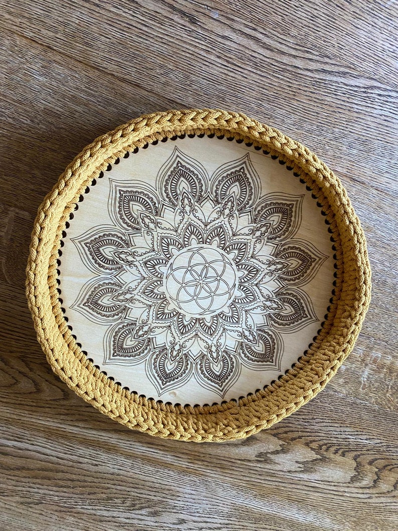 May include: A round wooden tray with a crocheted mustard yellow border. The tray features a laser-etched mandala design with a flower of life in the center.