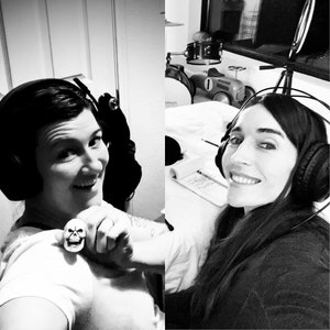 May include: Two people wearing headphones are seated at a table. One person is holding a skull ring. The other person is smiling and looking at the camera.