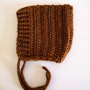 Peut inclure: Un bonnet de bébé en crochet marron avec une attache. Le bonnet est réalisé avec un motif de point simple et a un style classique.