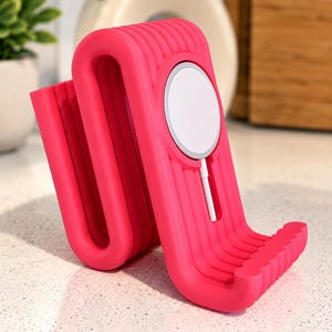 May include: A bright pink, ribbed, S-shaped phone stand featuring a circular white magnetic charging pad integrated into the front surface, with a white charging cable extending downward from the center of the circular magnetic component.