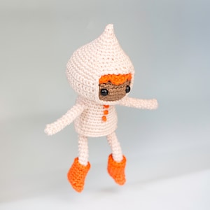 Puede incluir: Una muñeca de crochet con un abrigo rosa con capucha y botas naranjas. La muñeca tiene ojos marrones y pelo naranja.