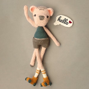 May include: A handmade crochet doll in the shape of a bear, wearing a teal top, gray shorts, and yellow roller skates. The doll has a speech bubble that says "hello" with a red heart. The bear is pink, gray, and white.