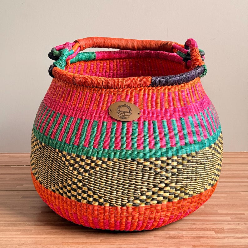 May include: A round, hand-woven basket featuring horizontal bands of vibrant orange, hot pink, teal, and black. The basket has a thick, wrapped handle and a central oval metal tag with the text Adiree Creations engraved on the front surface.