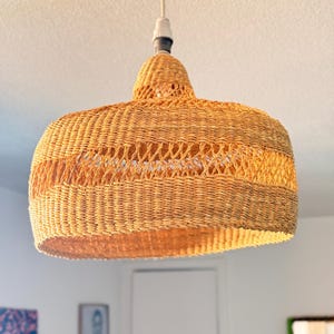 K&ouml;nnte beinhalten: Eine gewebte Pendelleuchte aus Rattan. Die Leuchte hat eine warme, nat&uuml;rliche br&auml;unliche Farbe und ein abgerundetes, glockenf&ouml;rmiges Design. Die Leuchte h&auml;ngt an einer wei&szlig;en Decke.