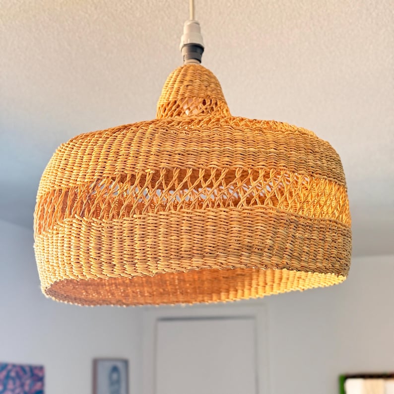 K&ouml;nnte beinhalten: Eine gewebte Pendelleuchte aus Rattan. Die Leuchte hat eine warme, nat&uuml;rliche br&auml;unliche Farbe und ein abgerundetes, glockenf&ouml;rmiges Design. Die Leuchte h&auml;ngt an einer wei&szlig;en Decke.