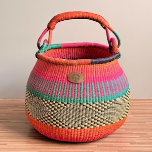 May include: A round, hand-woven basket featuring a vibrant color palette of orange, hot pink, teal, and black. The basket has a single, thick, orange-wrapped handle and a small, oval-shaped metal tag on the front with an engraved logo design.