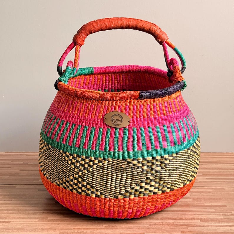 May include: A round, hand-woven basket featuring a vibrant color palette of orange, hot pink, teal, and black. The basket has a single, thick, orange-wrapped handle and a small, oval-shaped metal tag on the front with an engraved logo design.