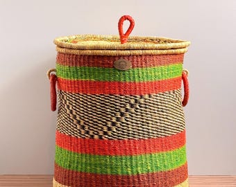 May include: A cylindrical woven basket with a lid featuring a central red loop handle. The basket has horizontal stripes in tan, bright green, and orange, with a central section of dark brown and tan woven texture and two red side handles.