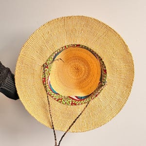 Puede incluir: Un sombrero de paja de ala ancha con una textura tejida en color tostado natural. El interior de la copa presenta un forro de tela colorido con un patr&oacute;n geom&eacute;trico rojo, verde y azul. Un cord&oacute;n trenzado de color marr&oacute;n oscuro est&aacute; sujeto al sombrero.