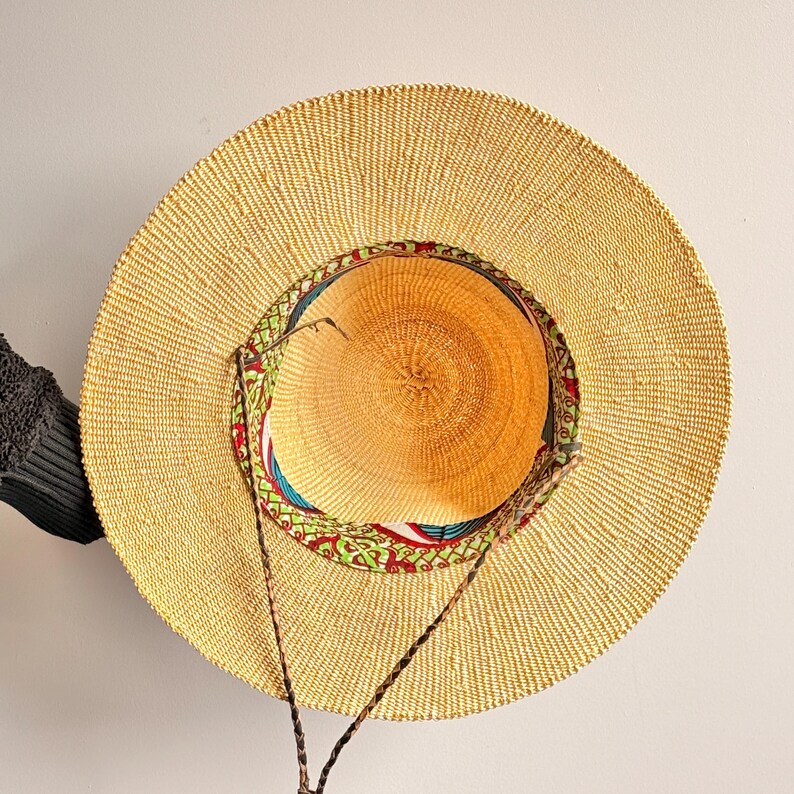 Puede incluir: Un sombrero de paja de ala ancha con una textura tejida en color tostado natural. El interior de la copa presenta un forro de tela colorido con un patr&oacute;n geom&eacute;trico rojo, verde y azul. Un cord&oacute;n trenzado de color marr&oacute;n oscuro est&aacute; sujeto al sombrero.