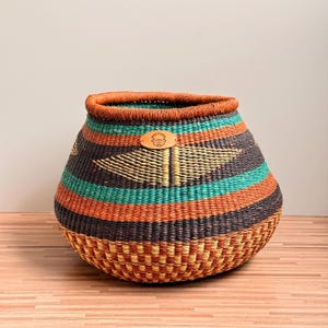 May include: A round, handwoven basket with a wide opening. It features horizontal stripes in teal, orange, and black, with a woven geometric design. A leather tag is attached to the front. The rim is trimmed with orange.