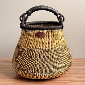 May include: A hand-woven basket featuring a rounded base and a wide opening with a dark brown rim. The body displays a geometric pattern in alternating yellow and black fibers. A black wrapped handle is attached, and a small brown leather patch.