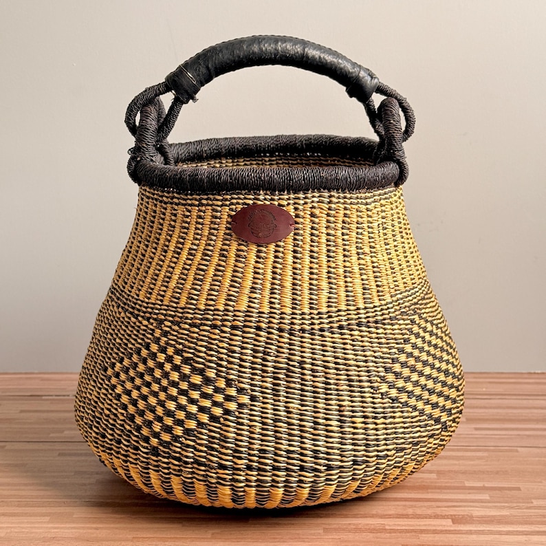 May include: A hand-woven basket featuring a rounded base and a wide opening with a dark brown rim. The body displays a geometric pattern in alternating yellow and black fibers. A black wrapped handle is attached, and a small brown leather patch.