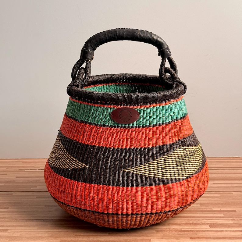 May include: A round woven basket featuring horizontal stripes in vibrant orange, black, and teal, with yellow triangular accents. The basket has a dark brown woven handle and a small, oval-shaped brown leather patch centered on the upper rim.