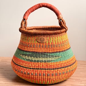 Può includere: Cesto intrecciato a mano con base arrotondata e manico. Il cesto presenta un motivo a strisce arancioni, verdi e nere. Il manico è avvolto in materiale marrone rossiccio e una piccola etichetta in pelle è attaccata al lato.